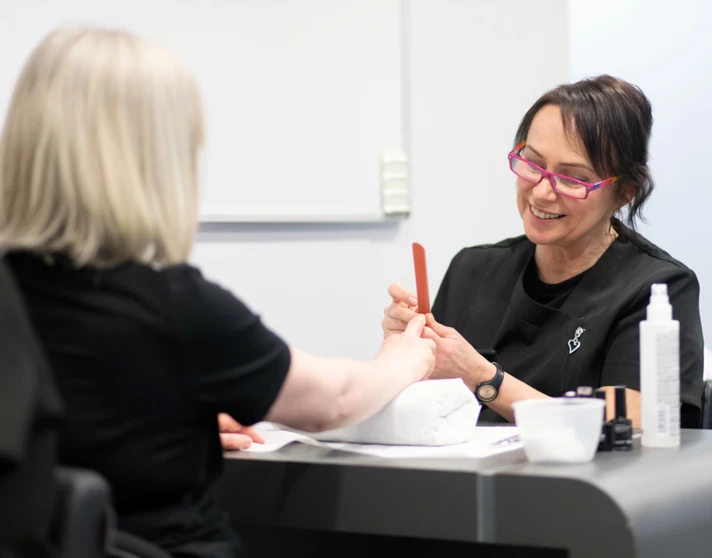 A beauty therapy student provides a manicure to a client, shaping nails with a file while smiling in a professional training setting. A beauty therapy student provides a manicure to a client, shaping nails with a file while smiling in a professional training setting.